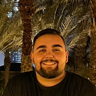 Habib Jaffer is standing and smiling on an outdoor balcony at night, surrounded by palm trees and lush greenery. The background features a beautifully lit pool area and the soft glow of distant city lights.